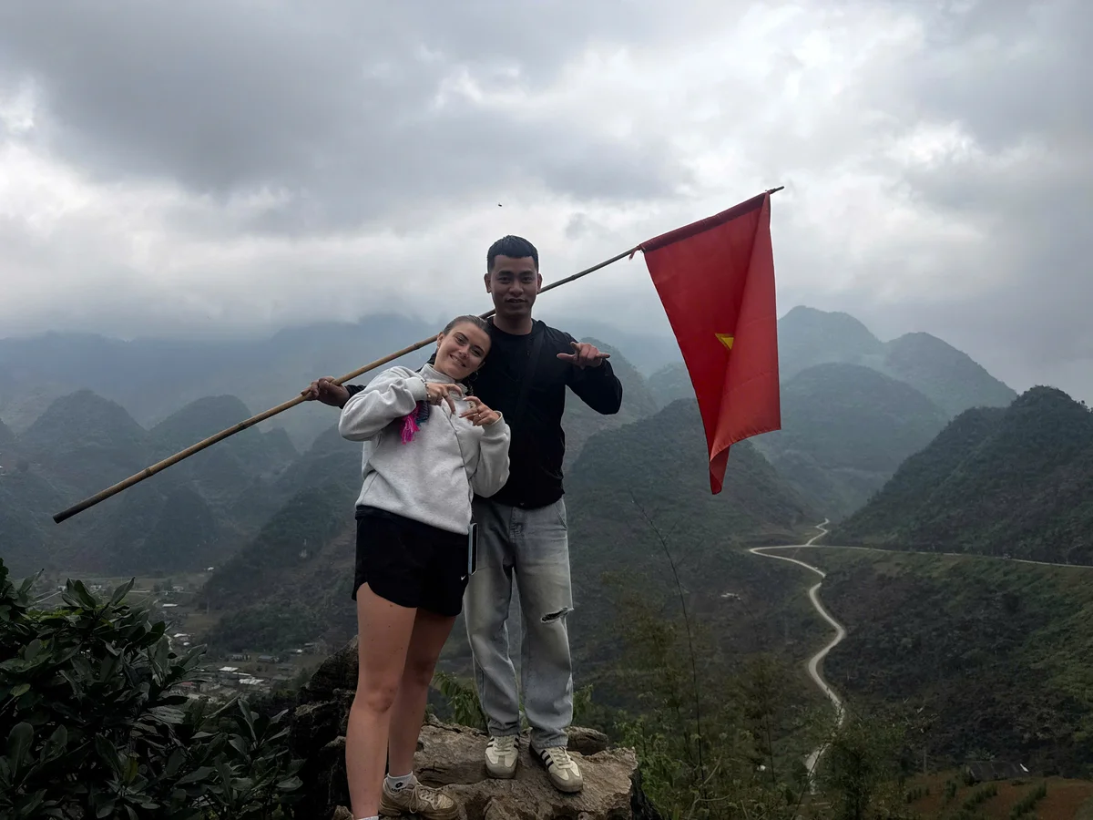 Two people pose with a red flag, there are mountains and small winding roads below