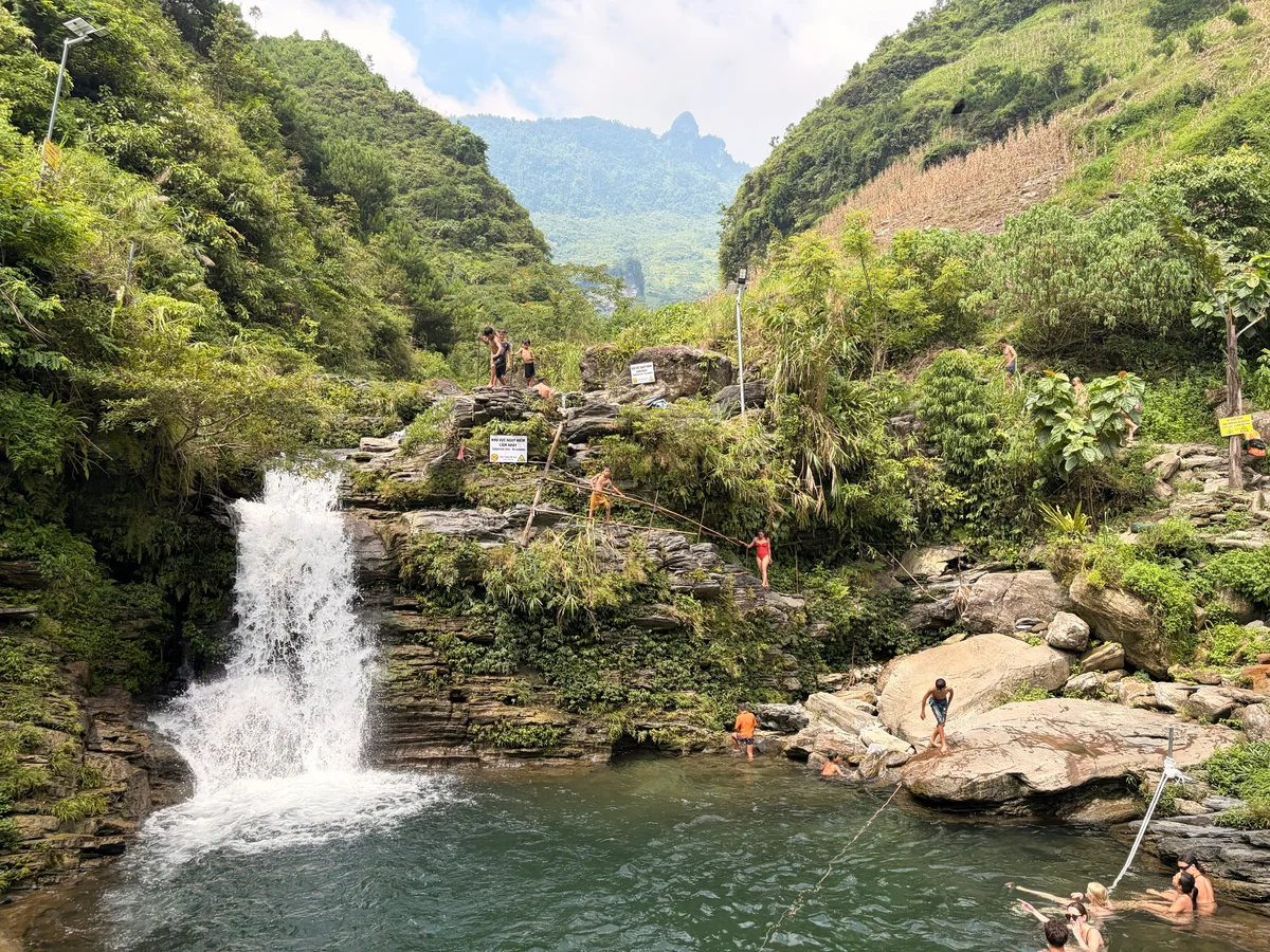 A waterfall falling into a pool of water with people around swimming and jumping
