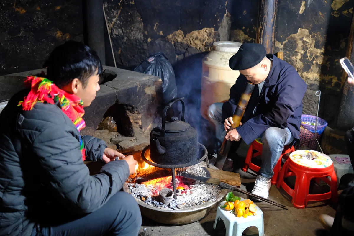 Two men in traditional Vietnamese clothing sit around a fire on small red stools