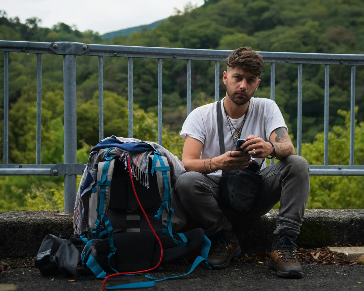 Man sits near a railing with forest behind on his phone with his big backpack next to him