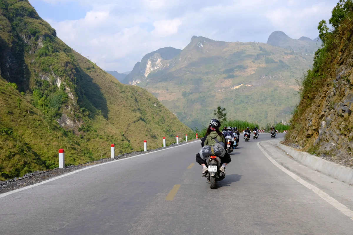 A group of motorbikes drive on a big wide road in a single line with mountains rising around them