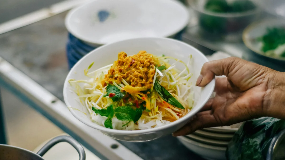 A hand delivers this bowl of food that looks like noodles, vegetables, and dried onions to a table