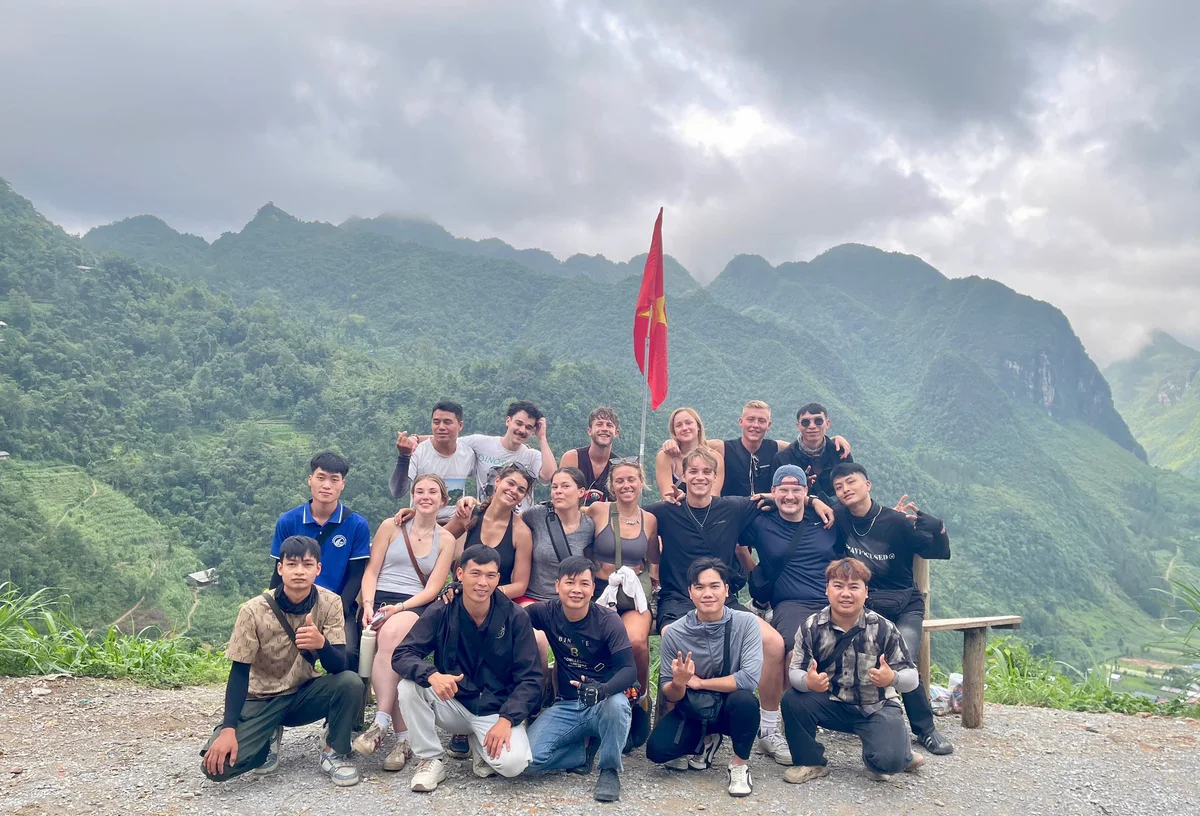 Group of people crouched and sitting posing for a photo with mountains behind and a red flag