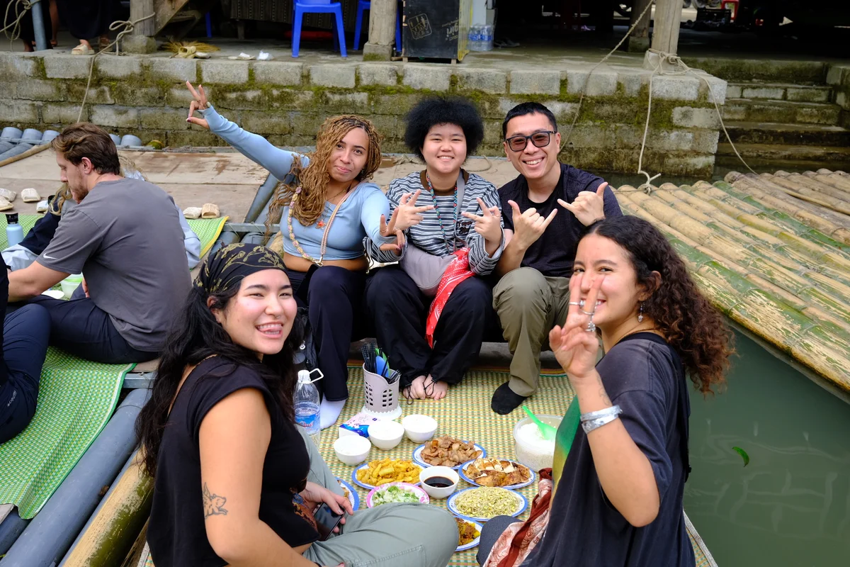 A group of people sit on a wooden bamboo raft with lunch laid out before them they smile and wave at the camera
