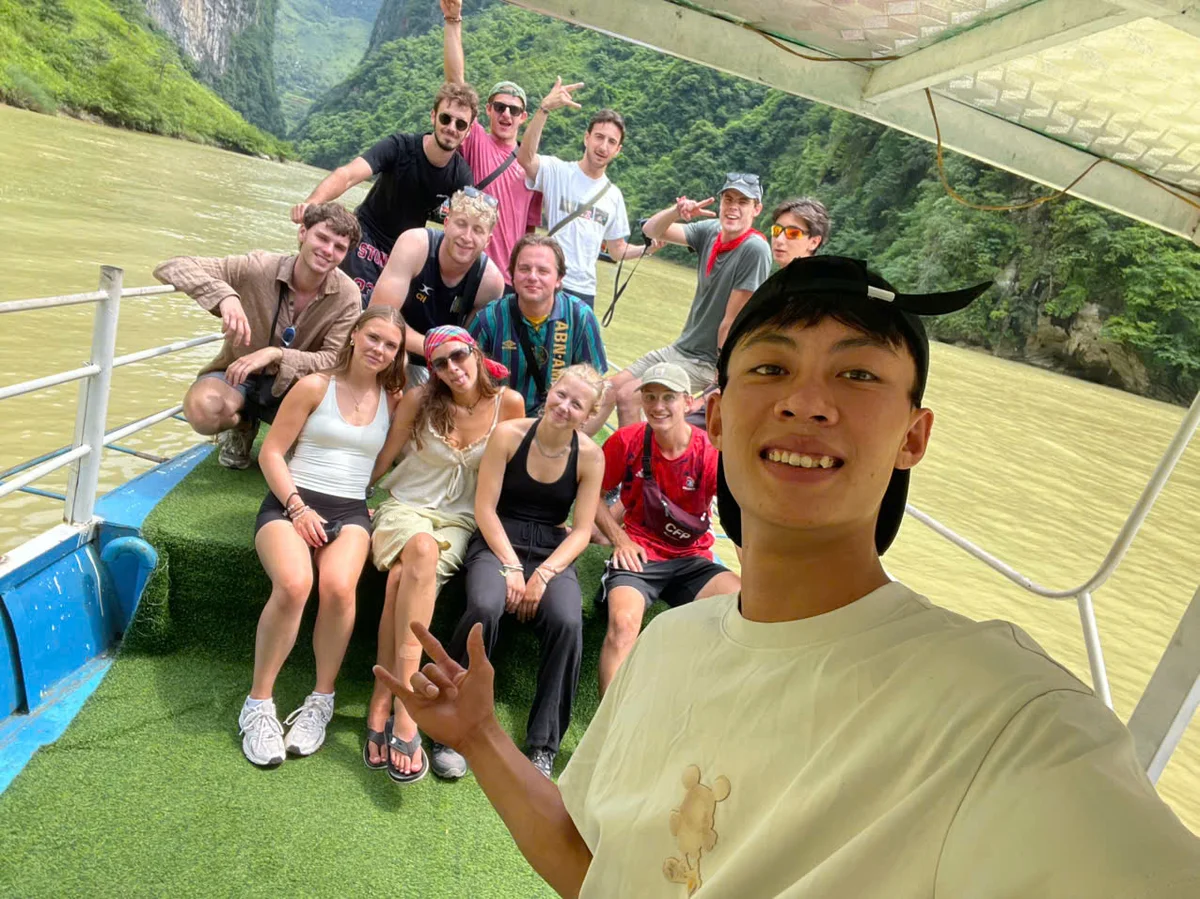 A man takes a selfie with a group of people smiling behind. The people sit on a boat that is making its way down the river.