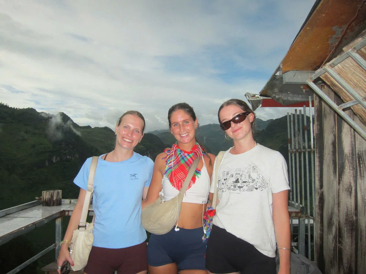 Three girls pose together with blue skies, mountains, and a homestay behind