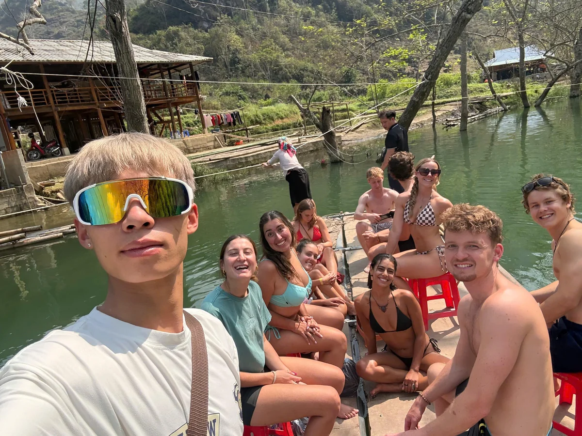 A man with yellow hair and sunglasses on takes  selfie with a group of people sitting on a wooden raft on a blue green river