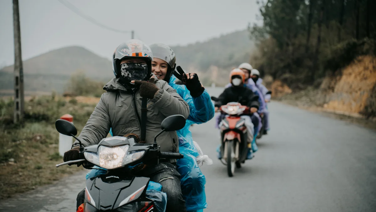 Two people on a motorbike smile and pose for the camera on a winding road with more motorbikes behind