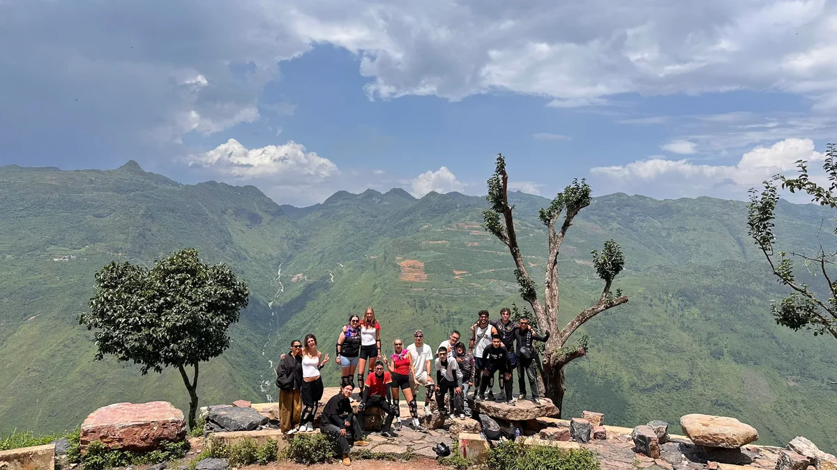 A group of people stand on the side of a road with mountain scenery behind and small trees