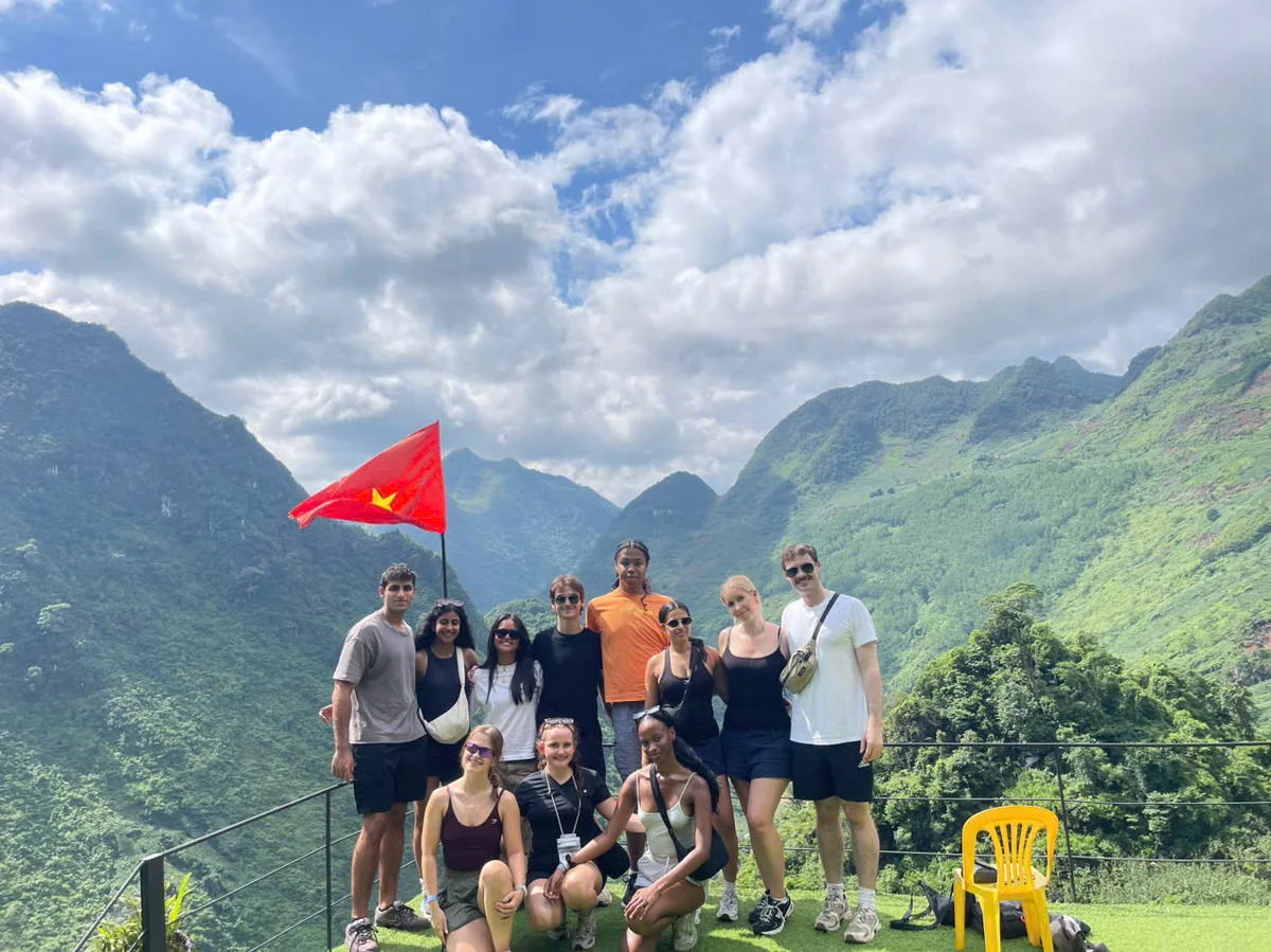 Group of people pose on a grassy bank in front of mountains with a red flag flying