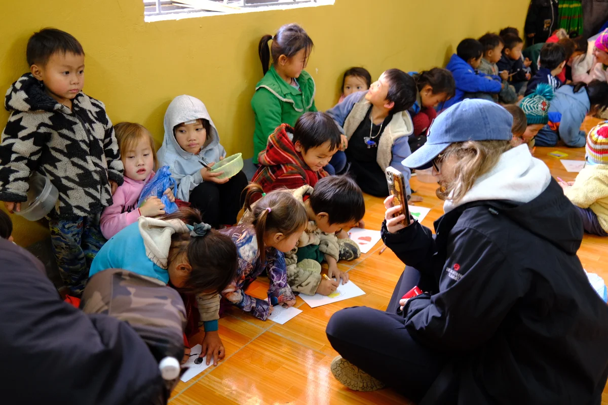 A group of children sit on the floor in colourful coats and jumpers while a woman with a blue hat and black jacket take a picture of them on her phone
