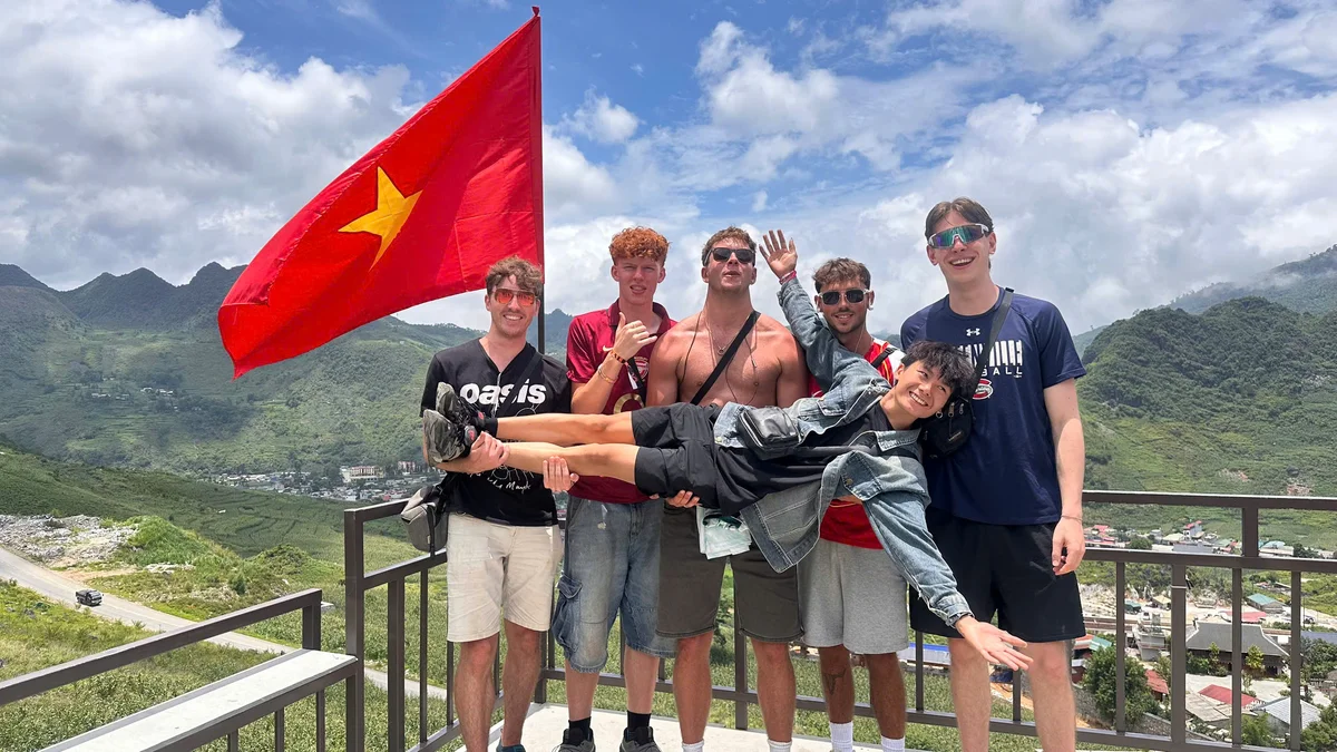A group of people hold up a Ha Giang Loop easy rider as they pose for a picture with a red flag with a yellow star on and mountains behind them