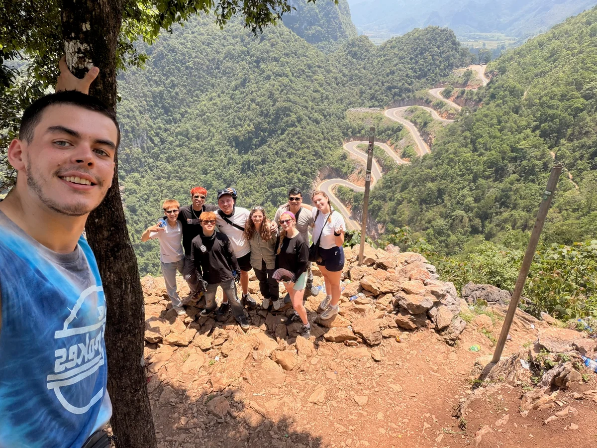 A man in a blue tshirt takes a selfie with a group of smiling people behind on a cliff edge in front of a winding mountain pass and green mountains