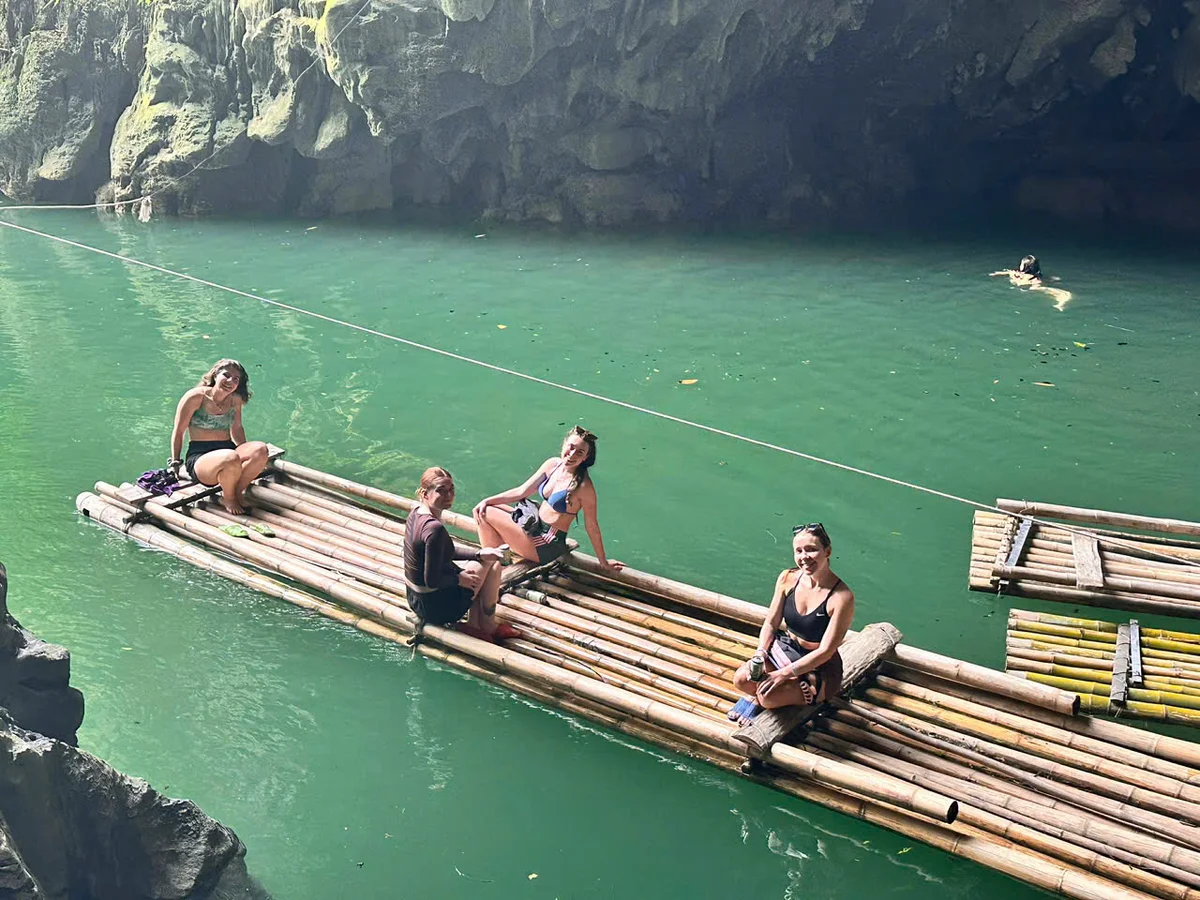 Four people sit on a bamboo raft on an emerald-green river in the sunshine