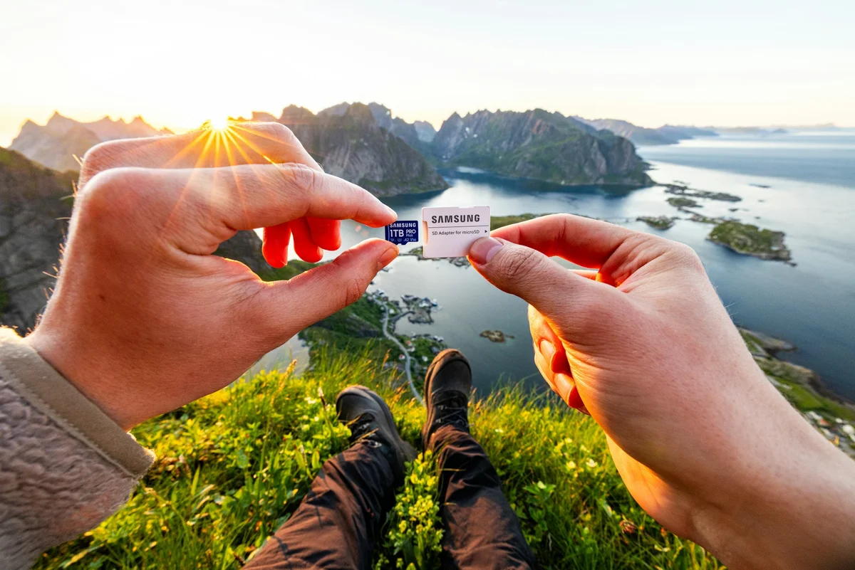 A pair of hands hold up a UK sim card with the word 'SAMSUNG' on it in front of a sunset and ocean and cliffs