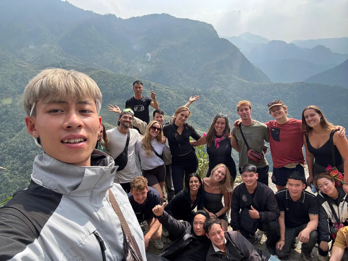 Man takes a selfie with group of people smiling and waving in front of mountains