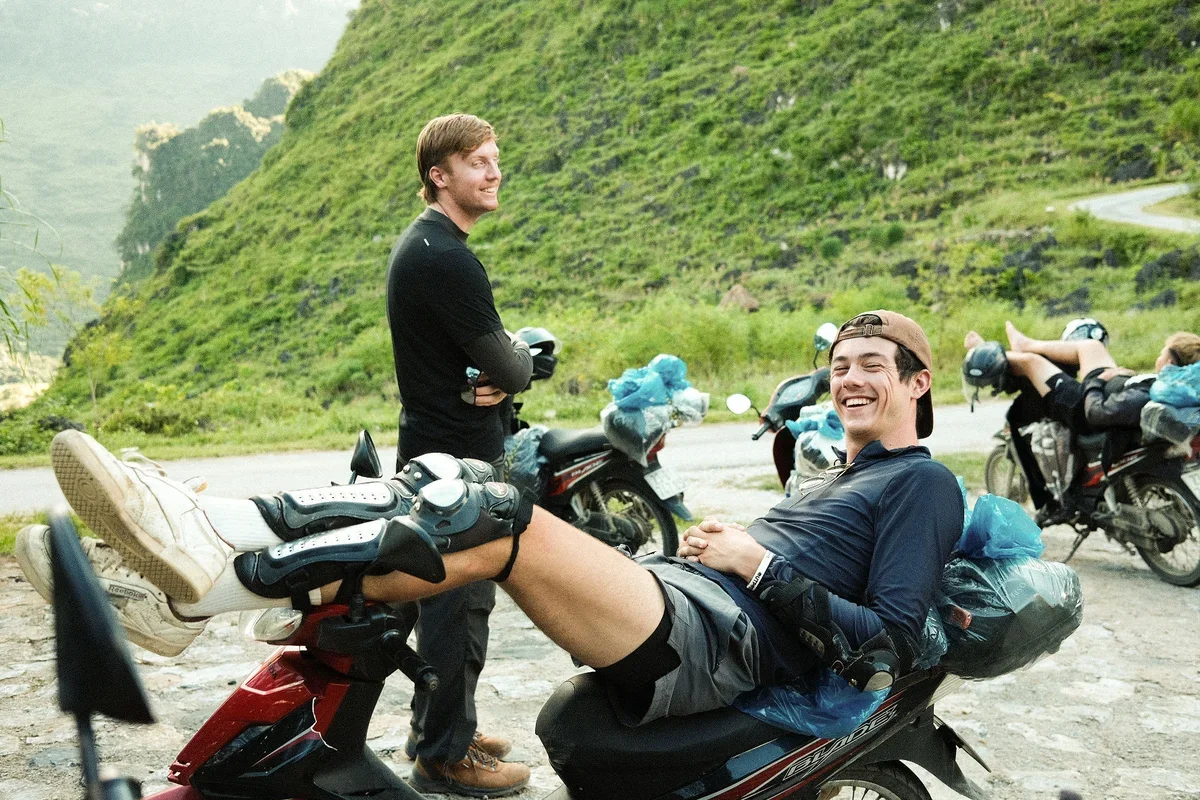 A man lies back on a motorbike with protective gear on and smiling with his legs up on the handlebars