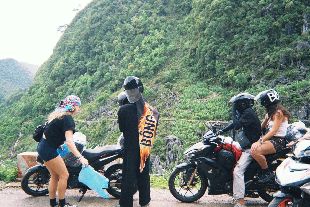 Two people sit on a motorbike, one person holds an orange flag that reads "Bong" and another girl prepares to get onto a motorbike with mountains behind