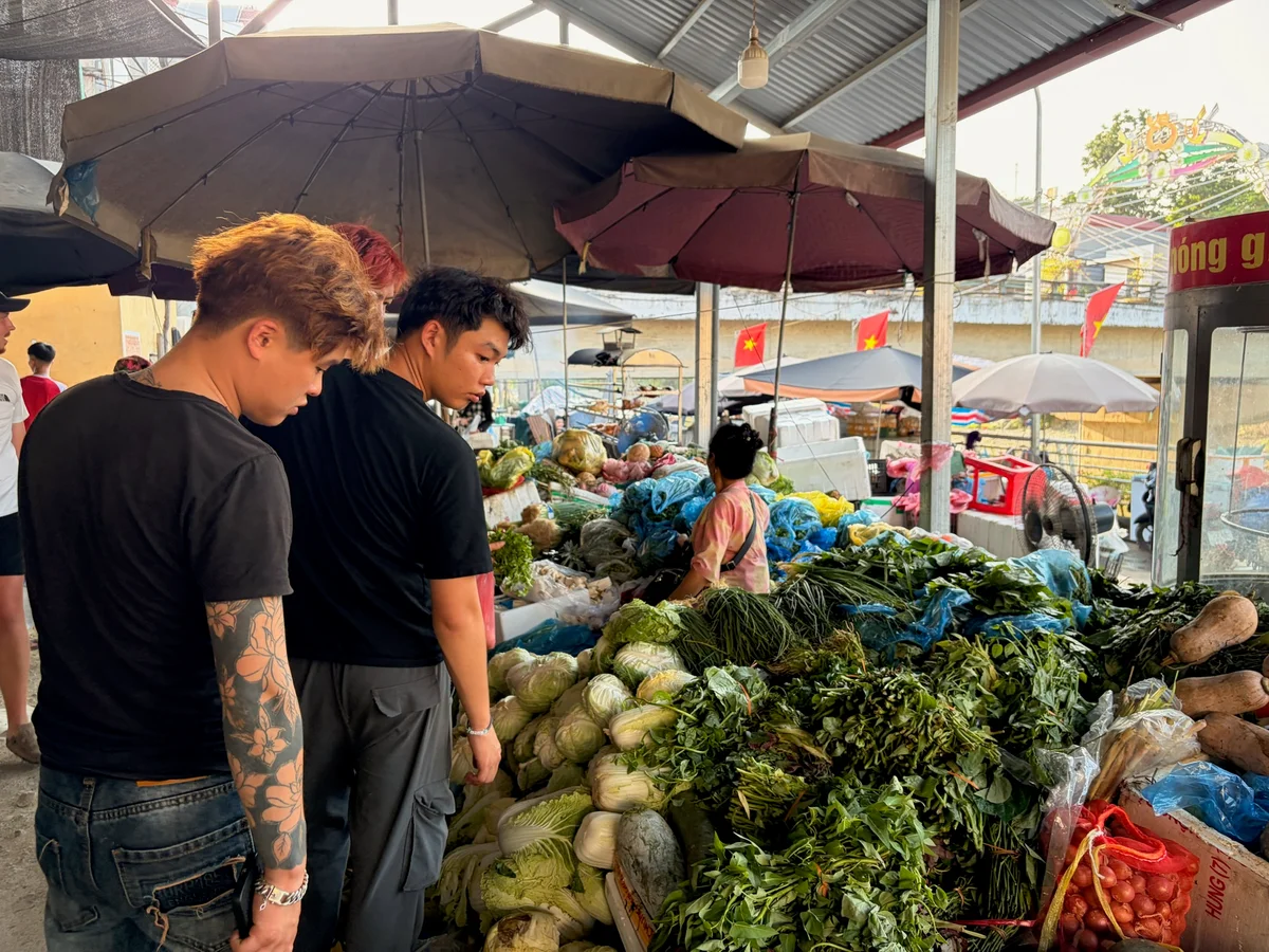 Two men look at vegetables at a store with a woman sitting taking money