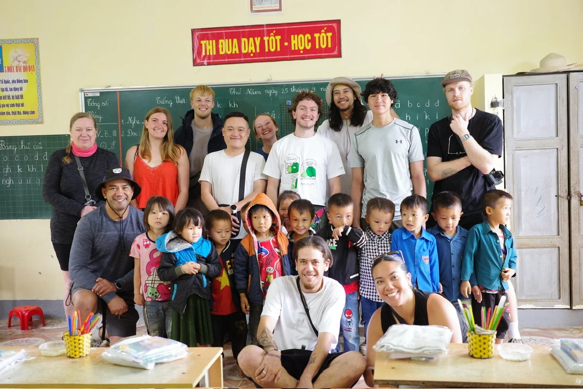 A group of people stand in front of a white board with desk around them and smile at the camera, there are children and older people