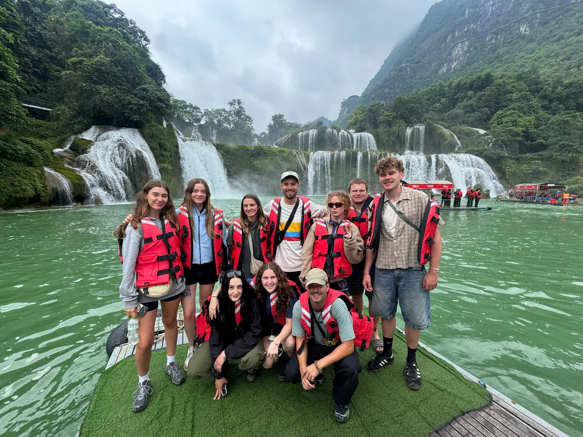 Group of people with life jackets on stand on a boat infront of a waterfall