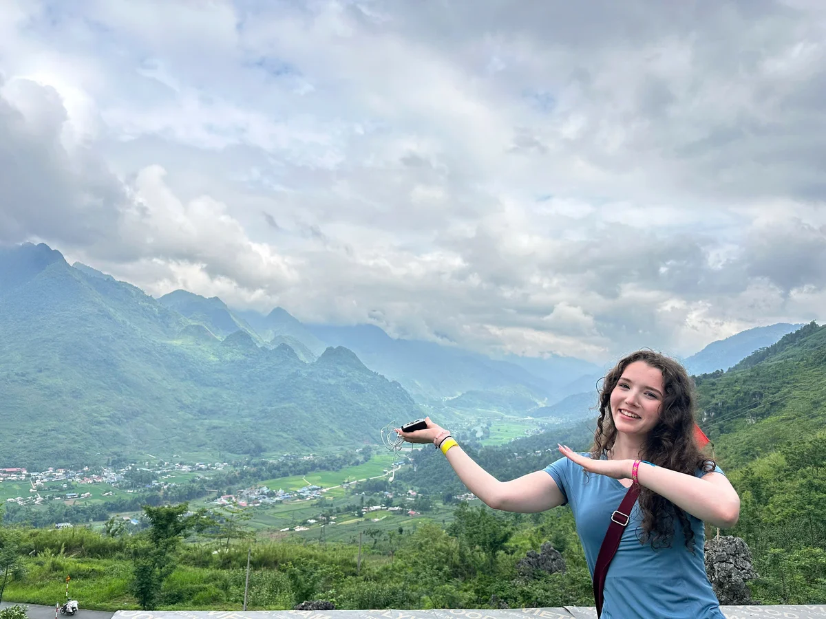 A woman stands in front of a mountain view with a valley below and holds her hands up to gesture to the view behind her