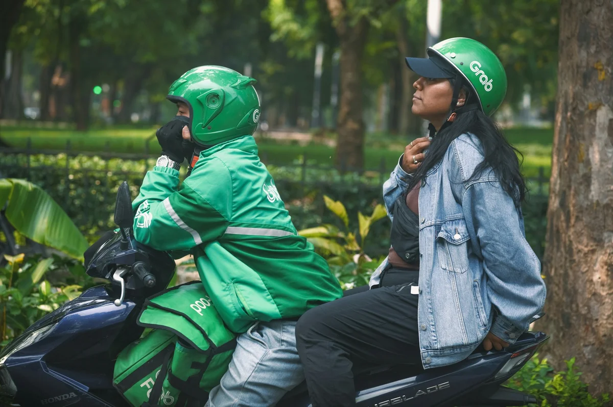 Woman sits behind a motorbike driver wearing a green Grab jacket