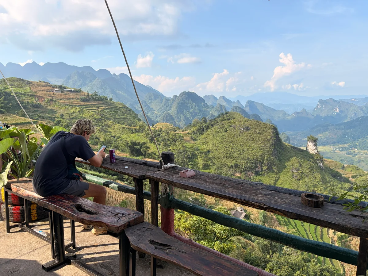 A man sits on his phone in front of an incredible view of mountains and blue skies