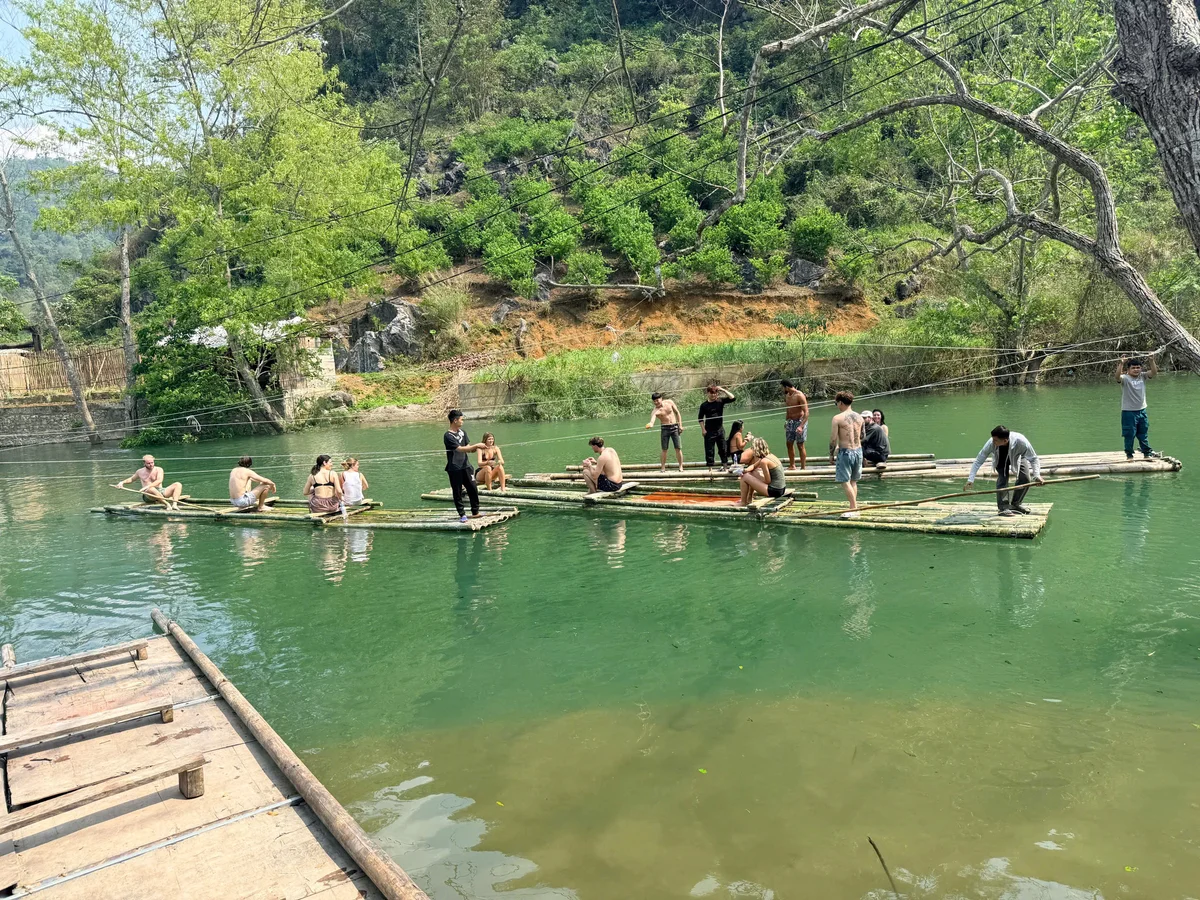 Several bamboo rafts carrying guests float down a green river with trees behind