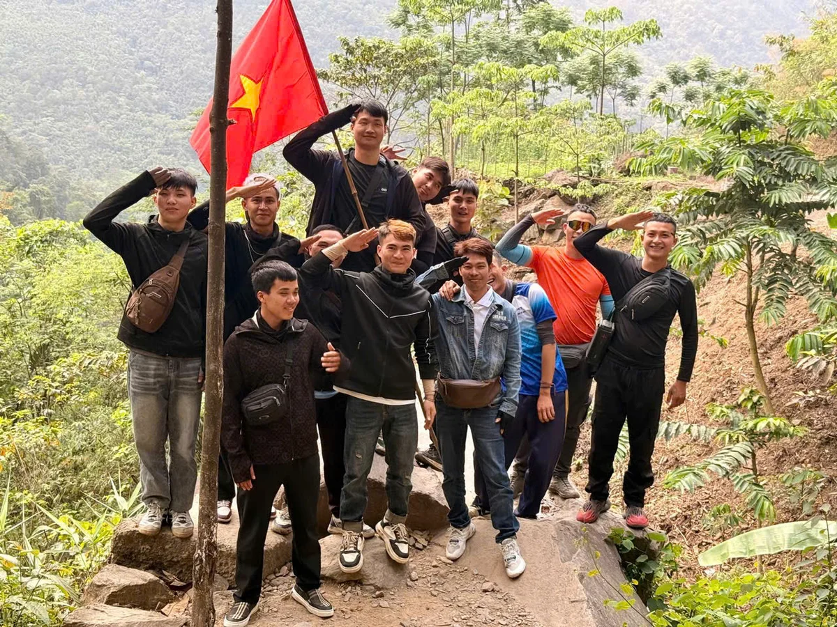Group of people stand by a post with a red flag attached to the top on a dirt track surrounded by trees