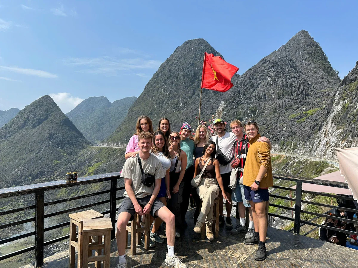 A group of people pose in front of a red flag with mountains and blue skies behind