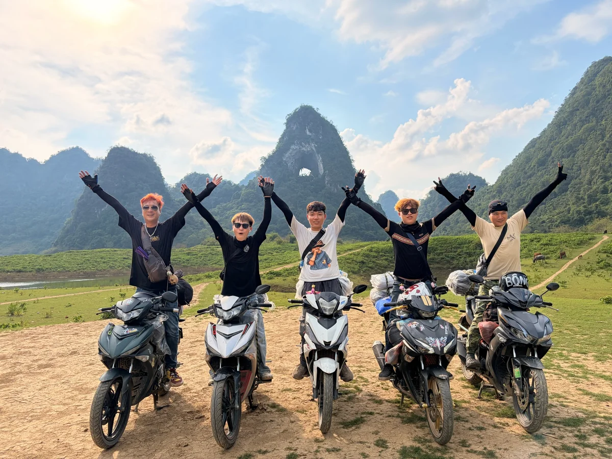 Group of 5 men stand on motorbikes in front of a mountain with a hole in it and throw their arms in the air