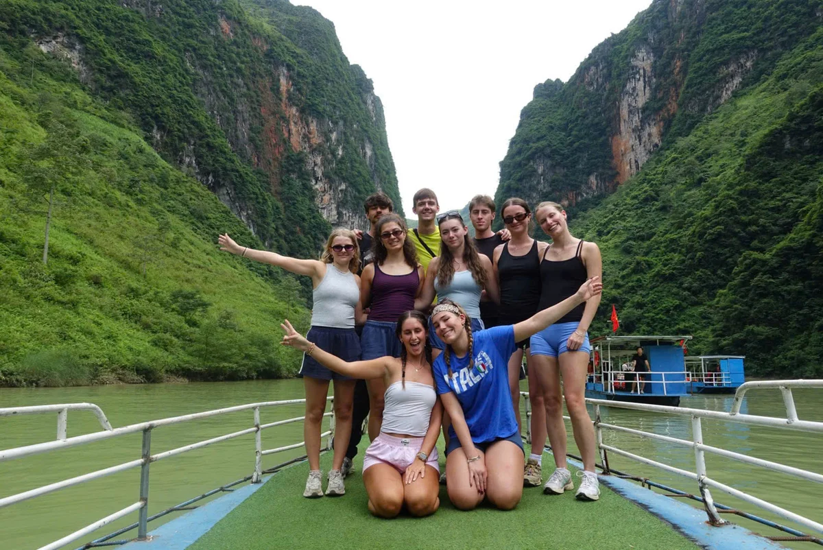 A group of people on a boat pose for the camera with a river and mountains behind