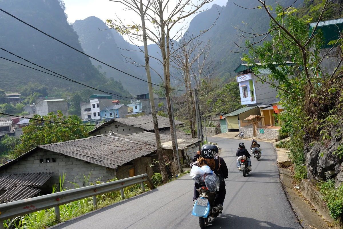 A group of people ride on motorbikes through mountains with houses and trees all around them