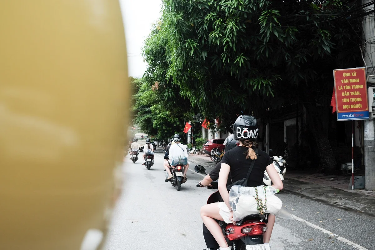 People ride motorbikes on a tree-lined street. A helmet with "BONG" is visible. Red signs in Vietnamese add vibrant color.