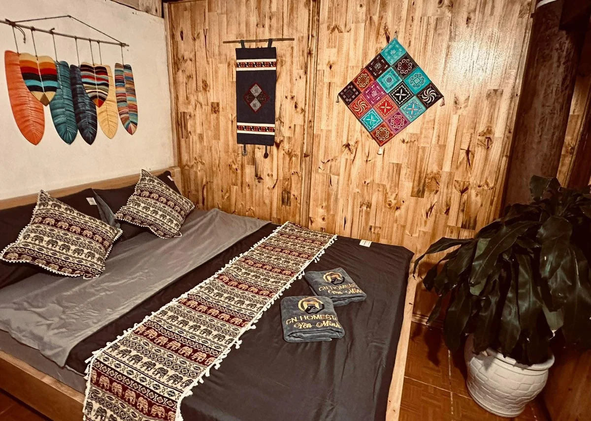 Cozy bedroom with wooden walls, colorful feather decor, patterned textiles on bed, and plant in the corner. Towels read "CN Homestay."