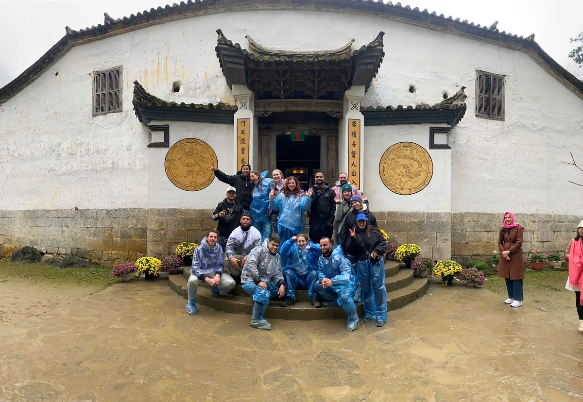 Group in blue raincoats smiles in front of a historic building with ornate details and Chinese characters. Cloudy weather, flowers lining steps.