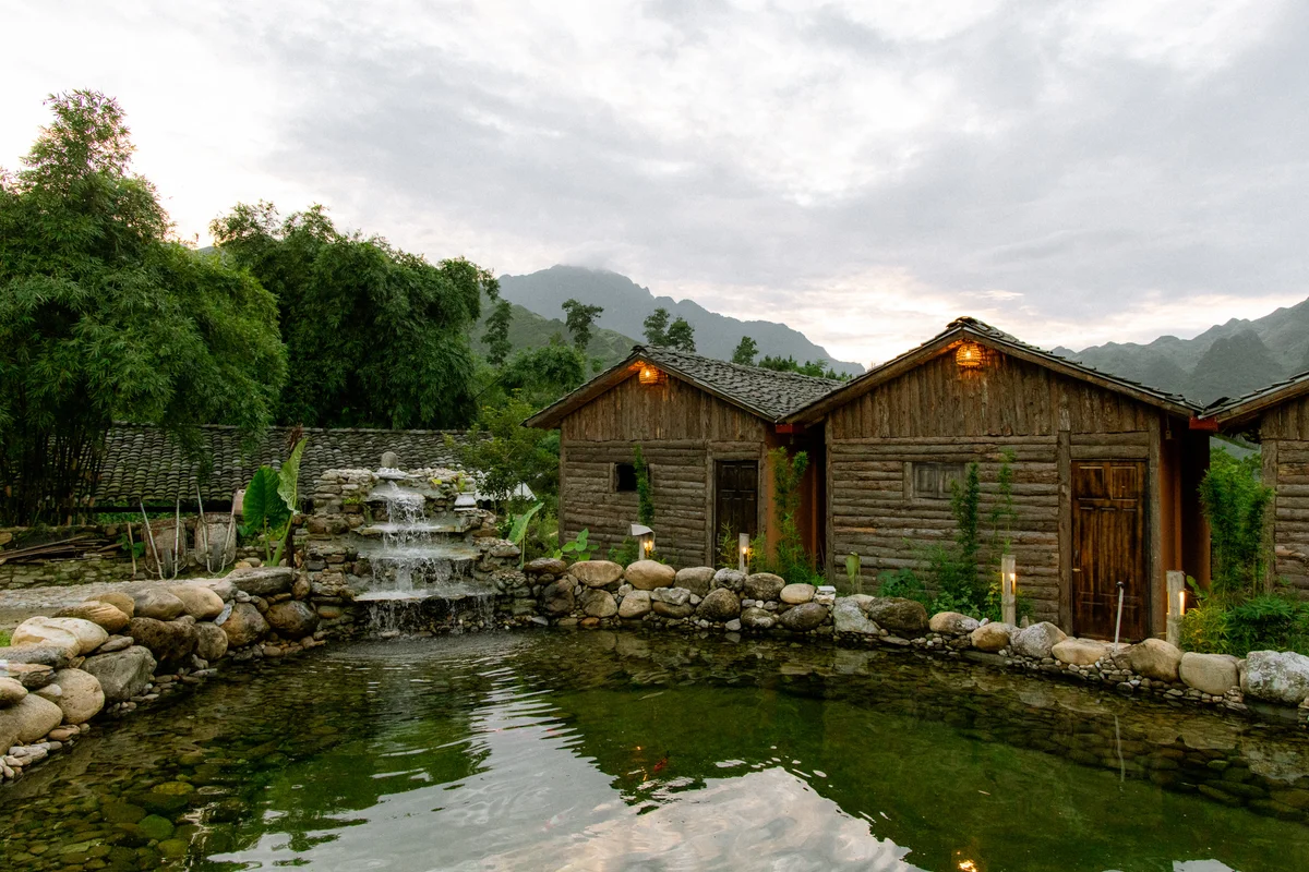 Wooden cabins by a pond beneath a cloudy sky, surrounded by lush green trees. A small waterfall flows over stacked rocks, creating a tranquil scenery.