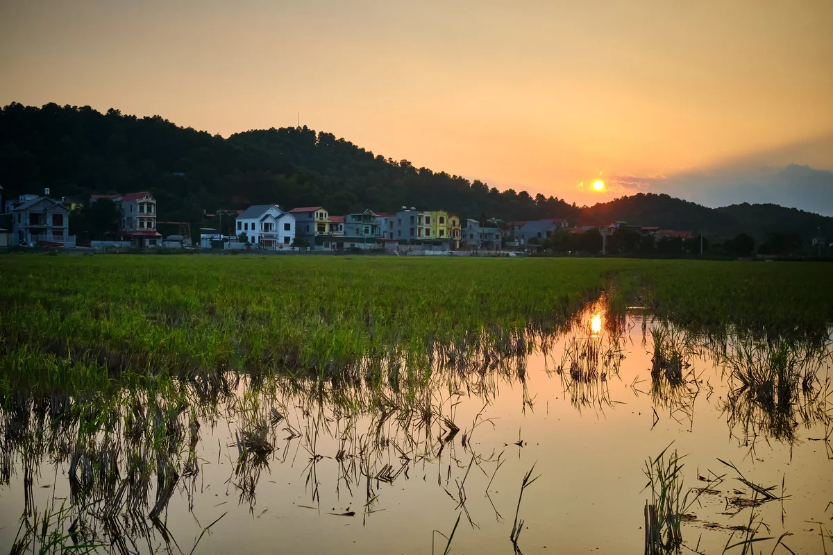 Sunset over a tranquil village with colorful houses, hills, and a reflective water-filled field in the foreground, creating a serene mood.