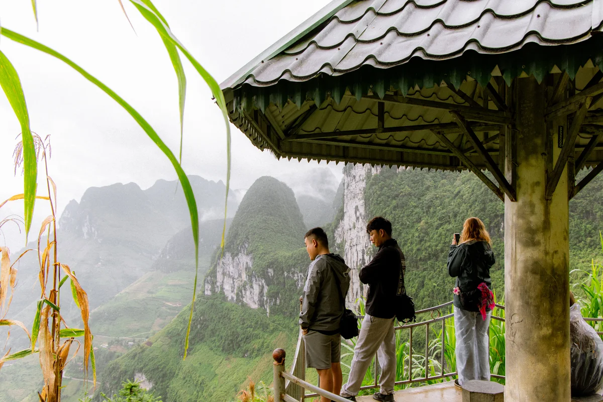 Three people stand under a gazebo, viewing misty green mountains. One takes a photo. Cornstalks are in the foreground.
