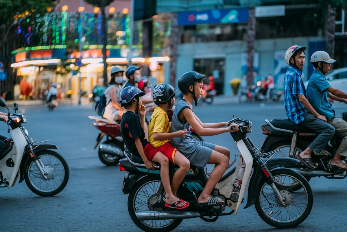 Three kids on a motorbike ride through a busy street, wearing helmets. Background shows illuminated city lights and other riders.