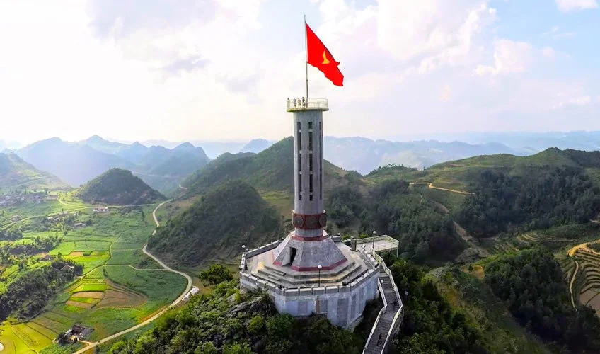 Tall tower with a red flag atop, set on a lush green hill with winding paths; mountainous background under a partly cloudy sky.