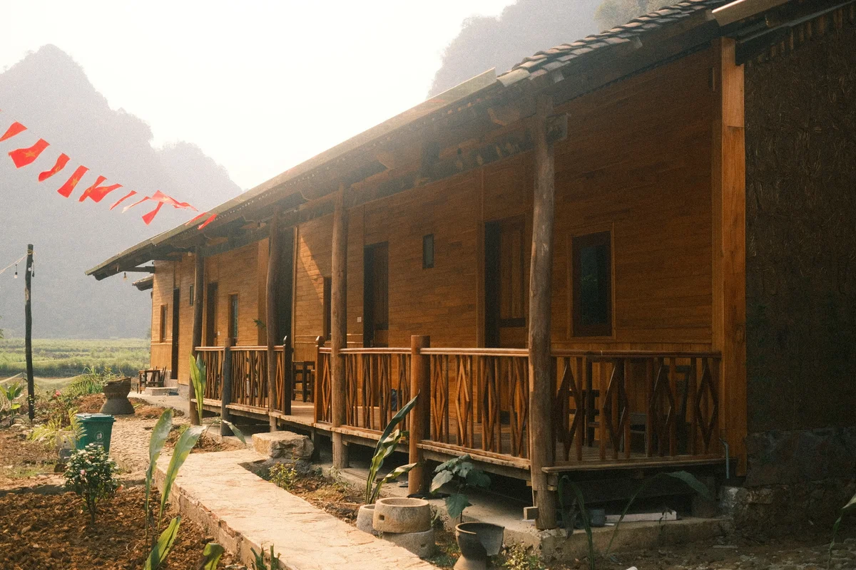 Wooden cabin with porch in a rural setting, red flags strung above. Mountains in the hazy background, lush plants, and a serene mood.