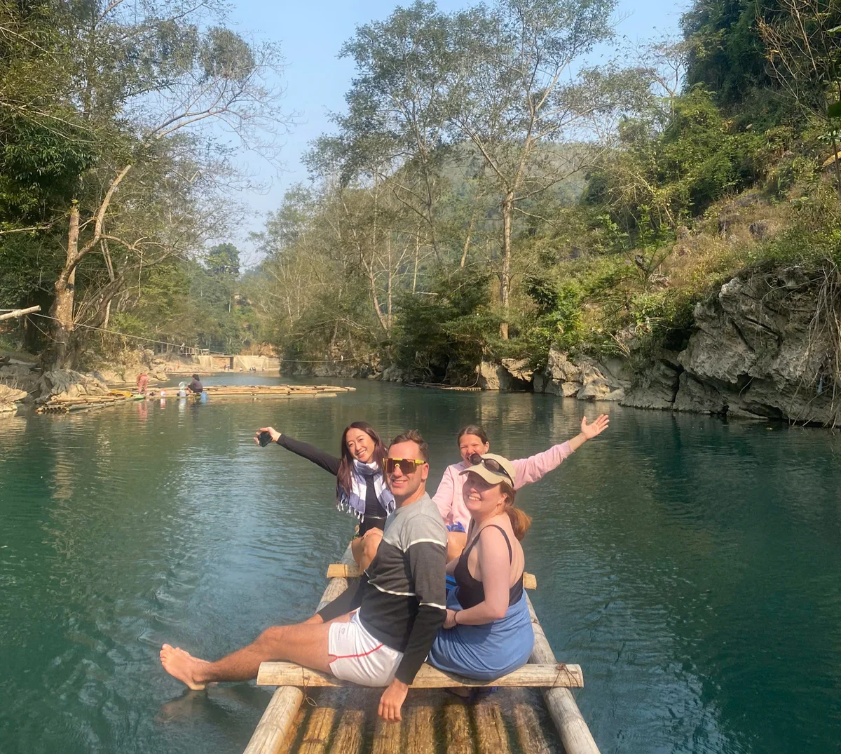 Four people smiling on a bamboo raft in a scenic river, surrounded by trees and rocky cliffs. Clear blue sky and calm water create a serene mood.