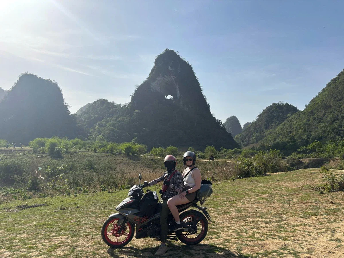 Two people on a motorcycle in a scenic landscape with lush green hills and a blue sky. They are wearing helmets and appear relaxed.