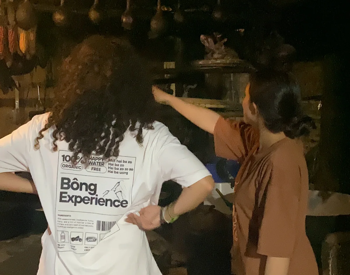 Two people in a dimly lit kitchen, one pointing at something. The back of a shirt reads "Bông Experience." Hanging tools and dark ambiance.