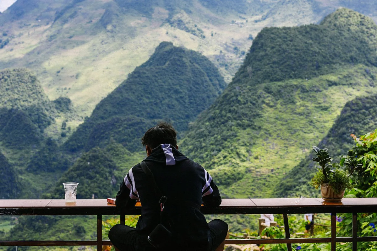A person sits at a table with a plant and cup, overlooking lush green mountains. The mood is peaceful, with vibrant greenery and rugged peaks.