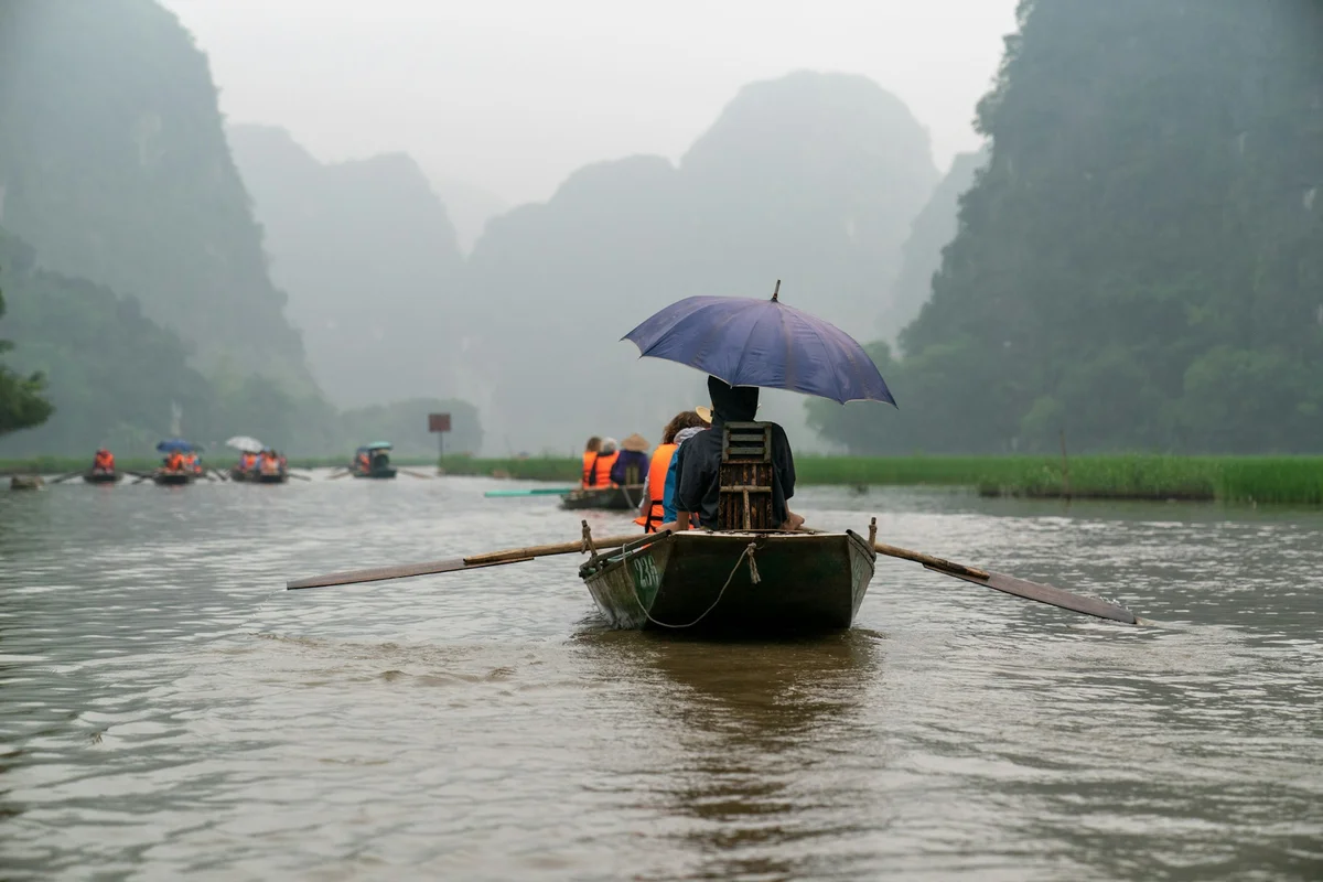 Boat on a misty river, person with blue umbrella rows. Passengers in life vests. Lush green hills in the background. Tranquil mood.