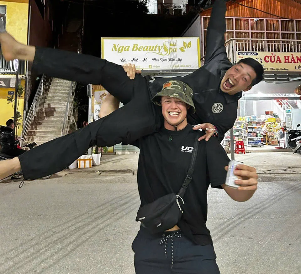 Two people in black clothes smiling, one lifts the other playfully in a city street. Background shows a spa sign and a store. Nighttime.