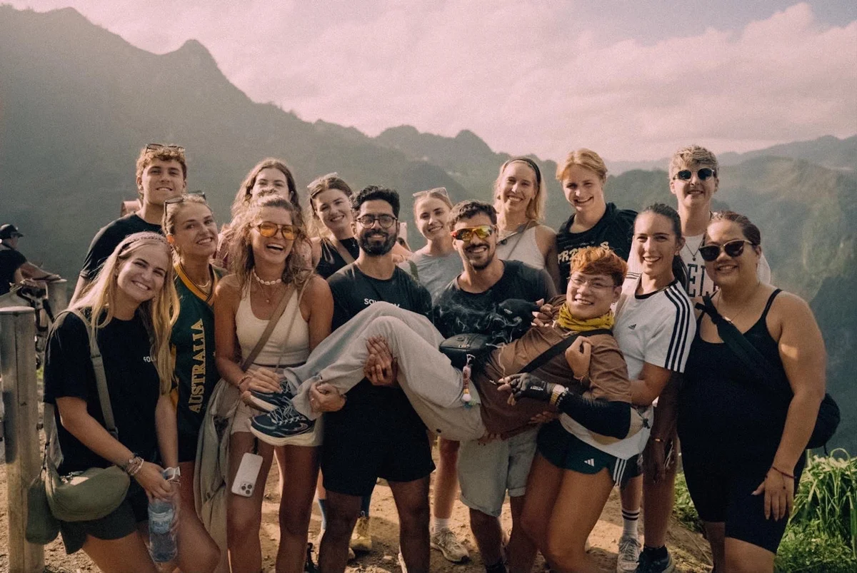 Group of smiling people pose on a mountain, one person is held up playfully. Text on shirt reads "Australia." Sunny, joyful setting.